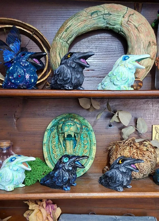 A collection of colorful handmade resin raven statues displayed on rustic wooden shelves with gothic home decor ornaments and fantasy props.